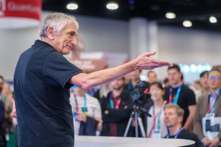 Attendees wearing lanyards gather to watch a speaker whose arm is extended as if presenting.